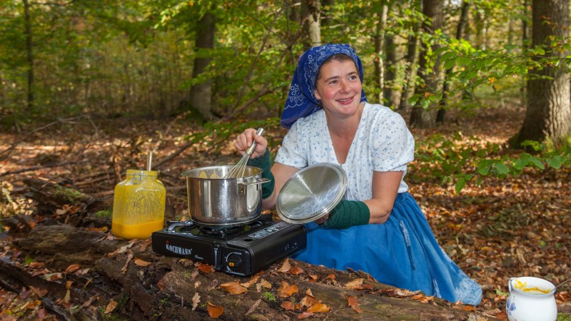 Fährfrau Yvonne die im Wald auf einem langen Baumstamm eine Suppe zubereitet